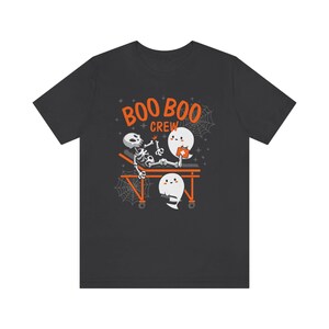 May include: Dark gray t-shirt with a Halloween-themed graphic. The design features a skeleton on a stretcher, with two ghost characters and the words "BOO BOO CREW" in orange. Spiderwebs and stars complete the design.