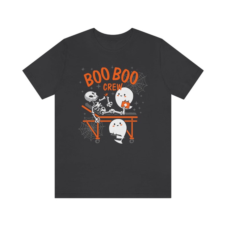 May include: Dark gray t-shirt with a Halloween-themed graphic. The design features a skeleton on a stretcher, with two ghost characters and the words "BOO BOO CREW" in orange. Spiderwebs and stars complete the design.