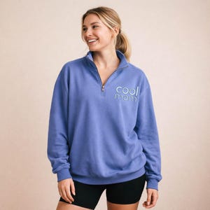 May include: A periwinkle blue pullover sweatshirt with a quarter-zip neckline. The text "cool mom" is embroidered in light blue on the left chest. The model is wearing black shorts. The background is a solid, neutral color.