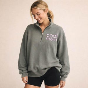 May include: A gray pullover sweatshirt with a quarter-zip neckline. The text "cool mom" is printed in a light purple color on the front. The model is wearing black shorts.
