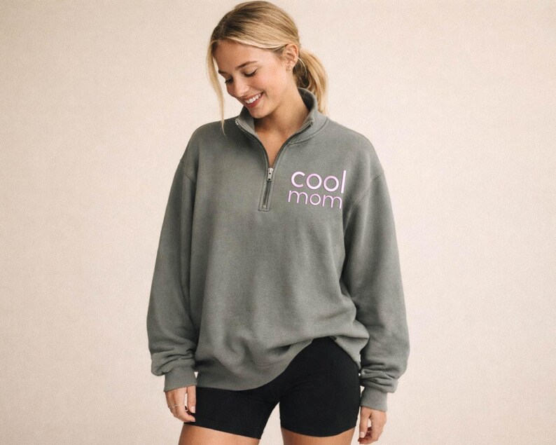 May include: A gray pullover sweatshirt with a quarter-zip neckline. The text "cool mom" is printed in a light purple color on the front. The model is wearing black shorts.