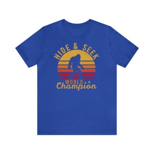 May include: A royal blue t-shirt with a vintage-style graphic of a Bigfoot walking in front of a sunset. The text reads "Hide & Seek World Champion".