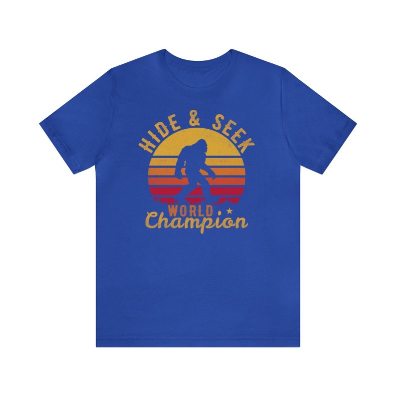 May include: A royal blue t-shirt with a vintage-style graphic of a Bigfoot walking in front of a sunset. The text reads "Hide & Seek World Champion".