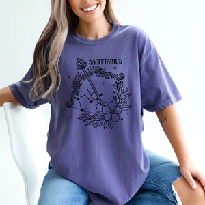 May include: A purple t-shirt featuring a black graphic of a Sagittarius zodiac sign with an arrow and flowers. The text "Sagittarius" is printed on the shirt.