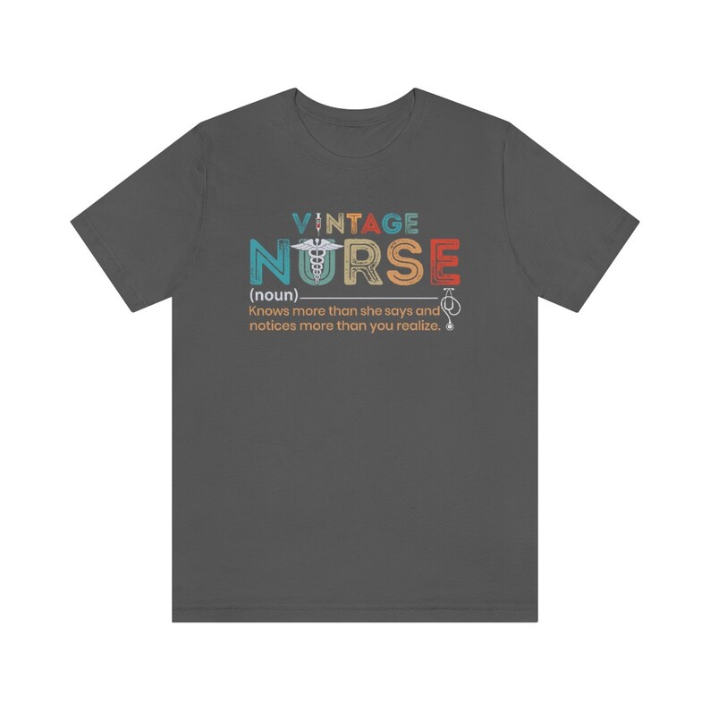 May include: A dark gray t-shirt with a vintage-style graphic that reads "VINTAGE NURSE (noun) Knows more than she says and notices more than you realize." The graphic features a colorful medical caduceus symbol.
