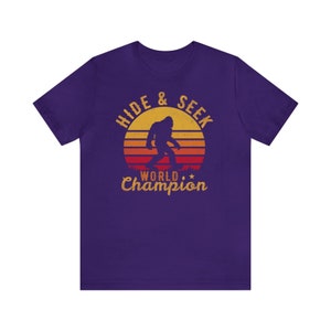May include: Purple t-shirt with a retro-style graphic of a Bigfoot walking in front of a sunset. The graphic says "Hide & Seek World Champion".