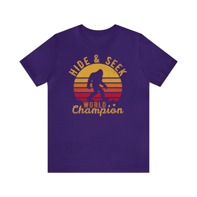 May include: Purple t-shirt with a retro-style graphic of a Bigfoot walking in front of a sunset. The graphic says "Hide & Seek World Champion".