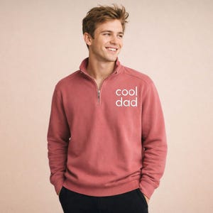 May include: A brick-red sweatshirt with a quarter-zip collar and the text "cool dad" in white. The sweatshirt has long sleeves and a relaxed fit. The model is wearing black pants.