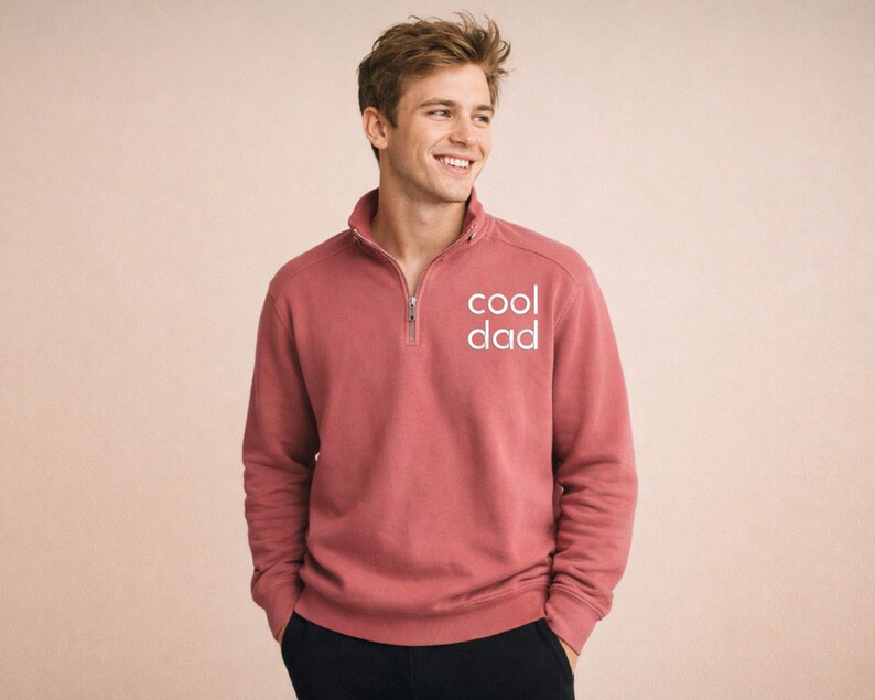 May include: A brick-red sweatshirt with a quarter-zip collar and the text "cool dad" in white. The sweatshirt has long sleeves and a relaxed fit. The model is wearing black pants.