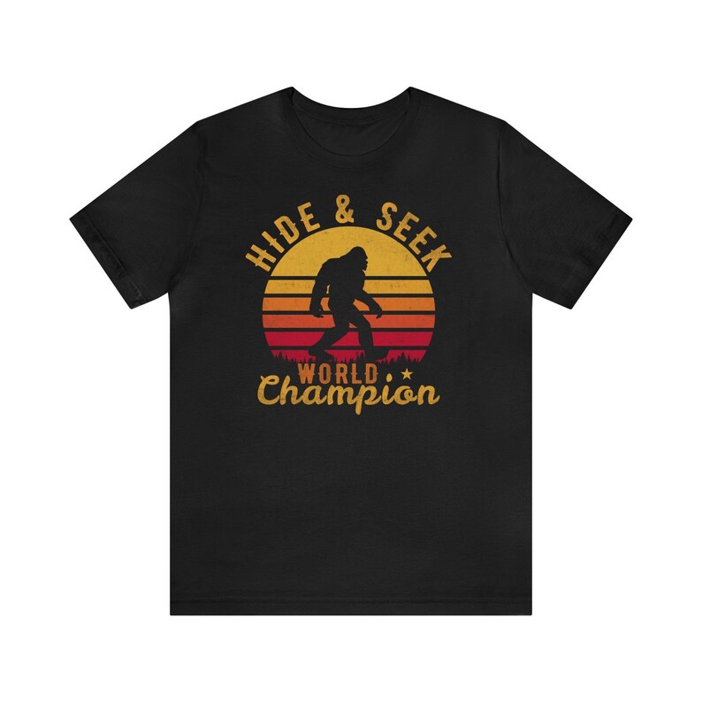 May include: Black t-shirt with a retro-style graphic of a Bigfoot walking in front of a sunset. The graphic says "Hide & Seek World Champion".