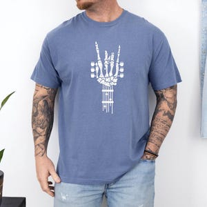 Pu&ograve; includere: Una maglietta blu con una grafica bianca di una mano scheletrica che fa il segno rock and roll. La mano tiene una chitarra.