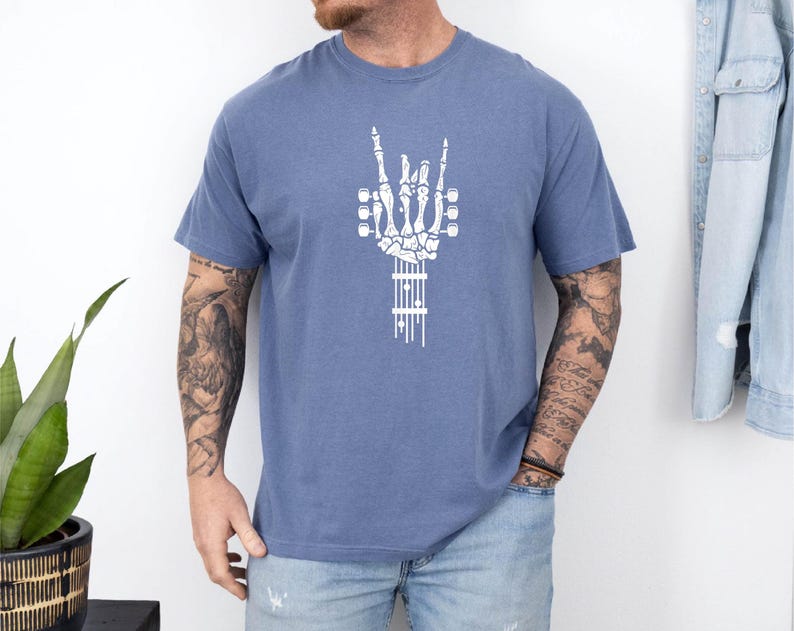 Pu&ograve; includere: Una maglietta blu con una grafica bianca di una mano scheletrica che fa il segno rock and roll. La mano tiene una chitarra.