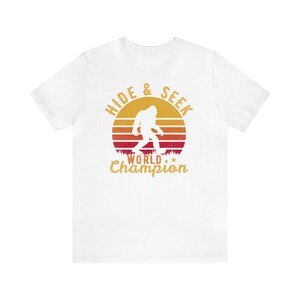 May include: White t-shirt with a retro sunset graphic featuring a silhouette of Bigfoot walking. The text "Hide & Seek World Champion" is printed in yellow below the graphic.