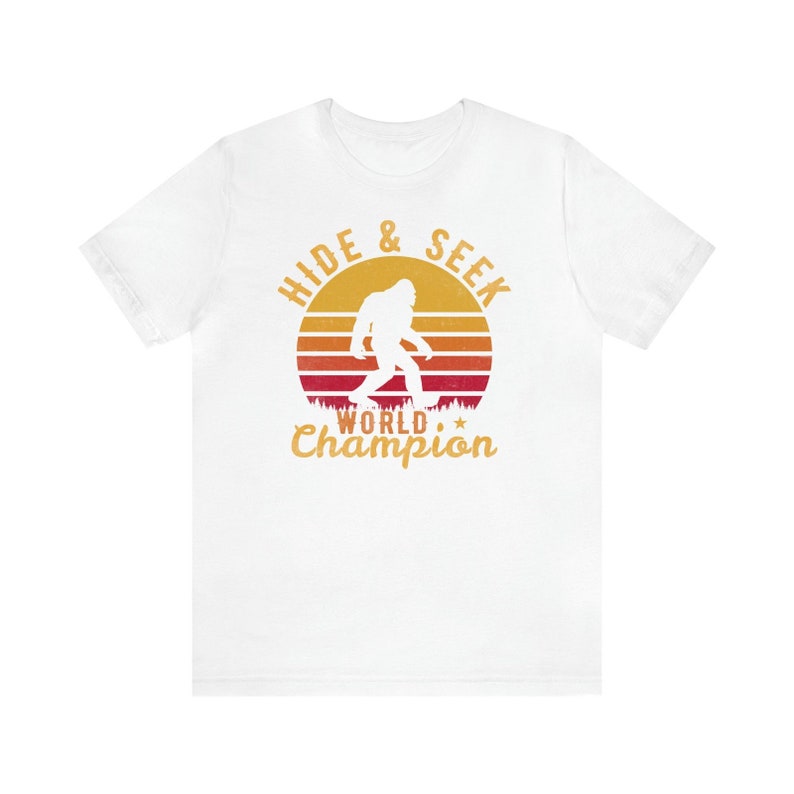 May include: White t-shirt with a retro sunset graphic featuring a silhouette of Bigfoot walking. The text "Hide & Seek World Champion" is printed in yellow below the graphic.