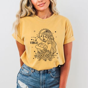 Peut inclure: Un t-shirt jaune avec une illustration en noir et blanc d'une femme portant une couronne de fleurs et tenant un globe. L'illustration est entour&eacute;e d'&eacute;toiles et le mot "VIRGO" est imprim&eacute; sous la femme.