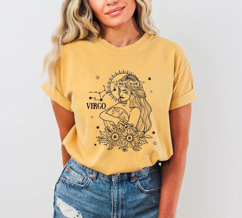 Peut inclure: Un t-shirt jaune avec une illustration en noir et blanc d'une femme portant une couronne de fleurs et tenant un globe. L'illustration est entour&eacute;e d'&eacute;toiles et le mot "VIRGO" est imprim&eacute; sous la femme.