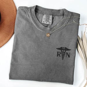 May include: A gray t-shirt with a black embroidered medical symbol and the letters "RN" on the left chest. The shirt is made of a soft, comfortable fabric.