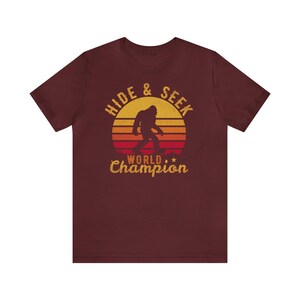 May include: A maroon t-shirt with a retro-style graphic of a Bigfoot walking in front of a sunset. The graphic says "Hide & Seek World Champion".