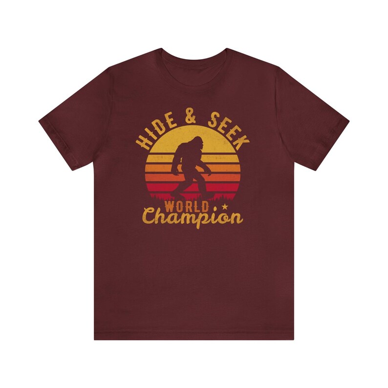 May include: A maroon t-shirt with a retro-style graphic of a Bigfoot walking in front of a sunset. The graphic says "Hide & Seek World Champion".