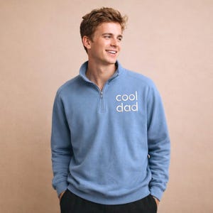 May include: A light blue pullover sweatshirt with a quarter-zip collar. The text "cool dad" is printed in white on the front. The sweatshirt has long sleeves and a relaxed fit.