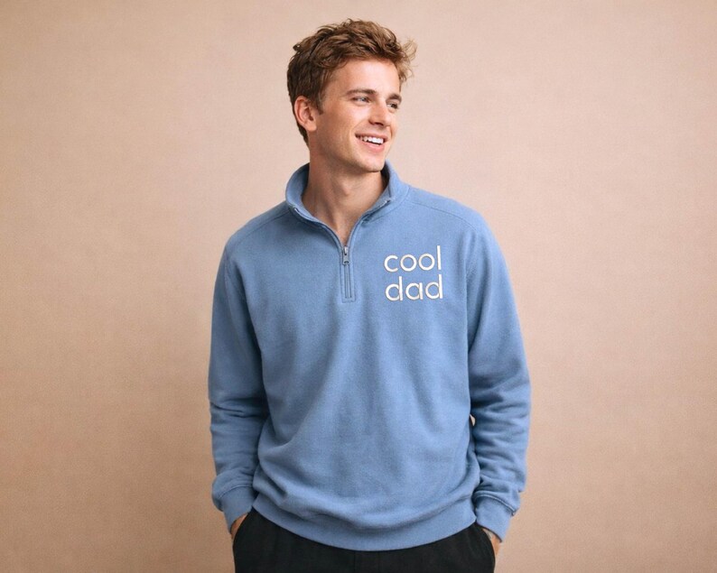 May include: A light blue pullover sweatshirt with a quarter-zip collar. The text "cool dad" is printed in white on the front. The sweatshirt has long sleeves and a relaxed fit.