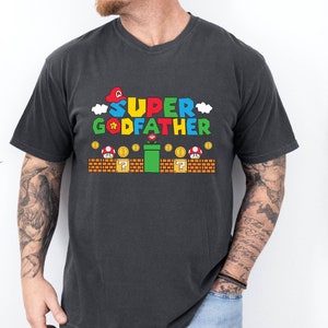 Puede incluir: Una camiseta negra con un gr&aacute;fico de un personaje de videojuego. El gr&aacute;fico dice "SUPER GODFATHER" en letras de colores con una estrella y nubes. El gr&aacute;fico tambi&eacute;n incluye una tuber&iacute;a verde, bloques de interrogaci&oacute;n y monedas.