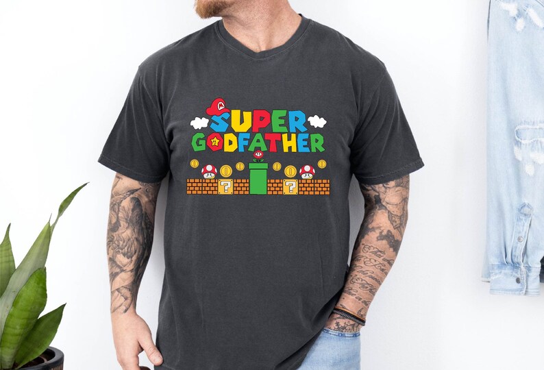 Puede incluir: Una camiseta negra con un gr&aacute;fico de un personaje de videojuego. El gr&aacute;fico dice "SUPER GODFATHER" en letras de colores con una estrella y nubes. El gr&aacute;fico tambi&eacute;n incluye una tuber&iacute;a verde, bloques de interrogaci&oacute;n y monedas.