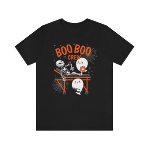 May include: Black t-shirt with a Halloween-themed graphic. The design features a skeleton on a stretcher attended by two ghosts, with the words "BOO BOO CREW" in orange. Spiderwebs and stars complete the design.