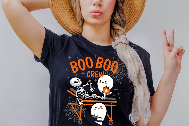 May include: Dark navy blue t-shirt with the words "BOO BOO CREW" in orange. The graphic features a skeleton on a stretcher being treated by a ghost, with two other ghosts and spiderwebs. A fun Halloween-themed design.