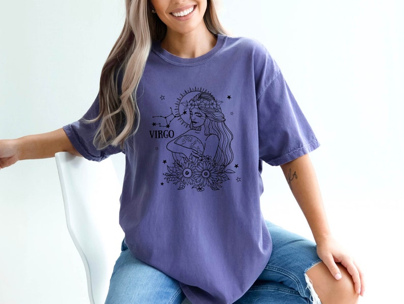 Peut inclure: Un t-shirt violet avec un motif noir et blanc d'une femme avec une couronne de fleurs tenant une lune et des &eacute;toiles. Le motif comprend &eacute;galement le mot "Virgo" et une constellation.