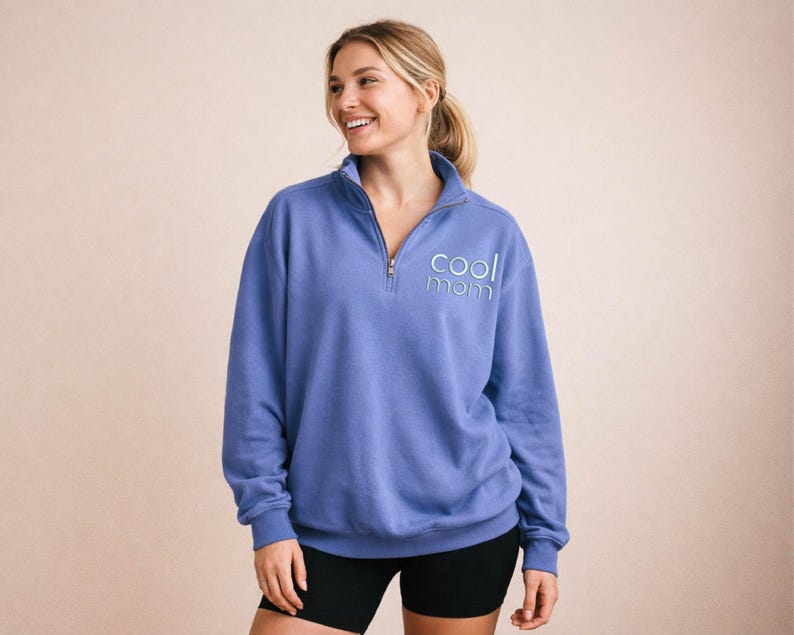 May include: A periwinkle blue pullover sweatshirt with a quarter-zip neckline. The text "cool mom" is embroidered in light blue on the left chest. The model is wearing black shorts. The sweatshirt has long sleeves and a relaxed fit.