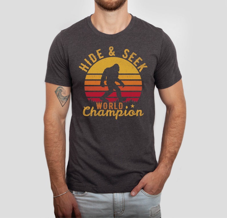 May include: A dark heather grey t-shirt with a retro-style graphic of a Bigfoot walking in front of a sunset. The text "Hide & Seek World Champion" is printed on the shirt.