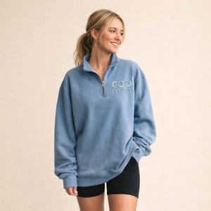 May include: A light blue sweatshirt with a quarter-zip neckline. The text "cool mom" is embroidered in light blue on the chest. The model is wearing black shorts.