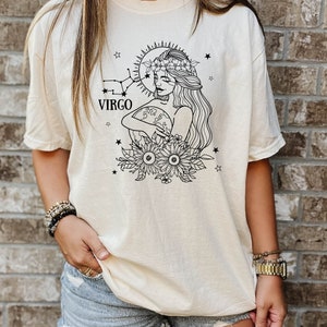 Peut inclure: Un t-shirt cr&egrave;me avec un motif noir et blanc d'une femme portant une couronne de fleurs et tenant un globe. Le motif comprend &eacute;galement des &eacute;toiles et des tournesols. Le texte "VIRGO" est imprim&eacute; sous la t&ecirc;te de la femme.