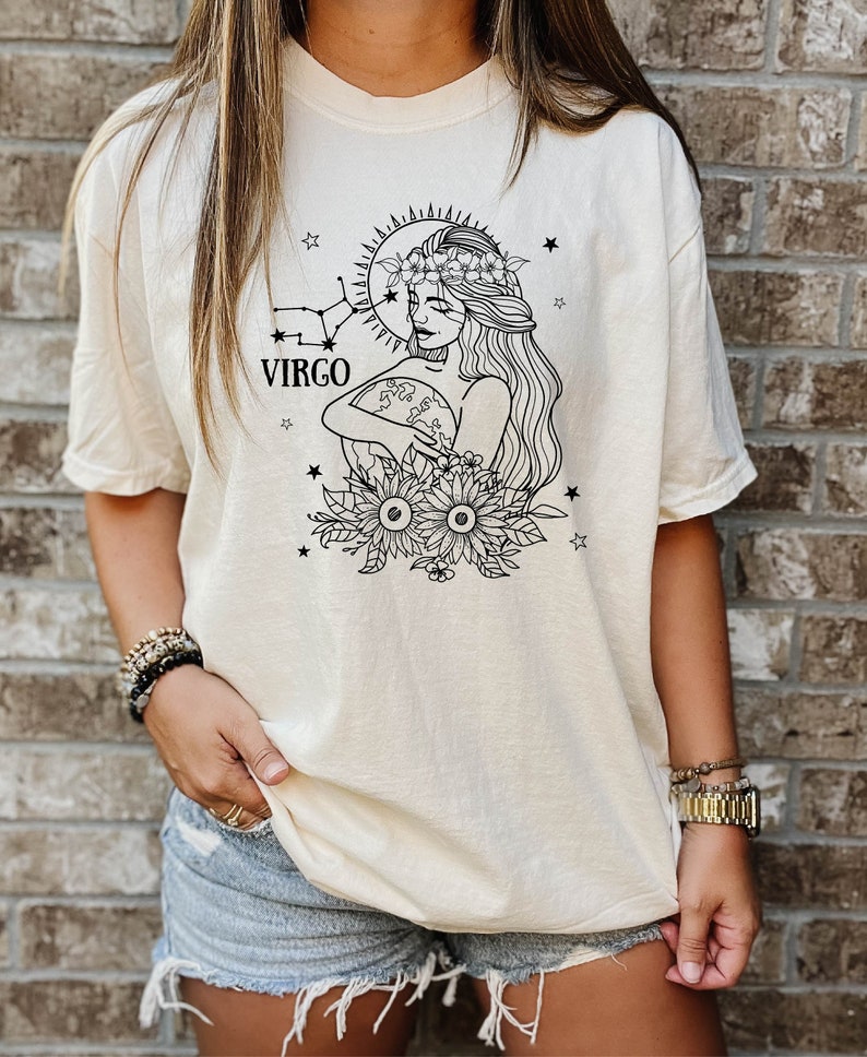 Peut inclure: Un t-shirt cr&egrave;me avec un motif noir et blanc d'une femme portant une couronne de fleurs et tenant un globe. Le motif comprend &eacute;galement des &eacute;toiles et des tournesols. Le texte "VIRGO" est imprim&eacute; sous la t&ecirc;te de la femme.