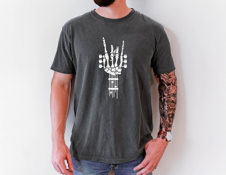 Pu&ograve; includere: Una maglietta grigia scura con una mano scheletrica bianca che fa il segno del rock and roll. La mano scheletrica tiene un manico di chitarra con le corde.