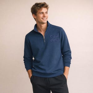 May include: A navy blue pullover sweatshirt with a quarter-zip collar and the embroidered text "cool dad". The long-sleeved top is made of a soft material and has a relaxed fit. The model is wearing black pants.