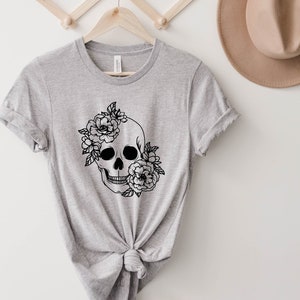 K&ouml;nnte beinhalten: Ein graues T-Shirt mit einer schwarz-wei&szlig;en Illustration eines Sch&auml;dels mit Blumen.