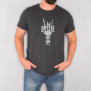 Pu&ograve; includere: Una t-shirt grigio scuro melange con una mano scheletrica bianca che fa il segno rock and roll, tenendo una chitarra. La chitarra &egrave; fatta di ossa e ha un contorno bianco.