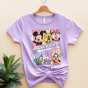 Puede incluir: Una camiseta de color lila claro con un dise&ntilde;o gr&aacute;fico de Mickey Mouse, Minnie Mouse, Donald Duck, Daisy Duck, Pluto y Goofy. El dise&ntilde;o gr&aacute;fico est&aacute; en un dise&ntilde;o rectangular con el texto "Mickey & Co. Est. 1928".