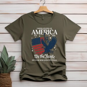 May include: An olive green t-shirt with a patriotic design. The graphic includes the text "UNITED STATES OF AMERICA" with the years 1776 and 2026, an American flag, a blue eagle, and the words "We the People".