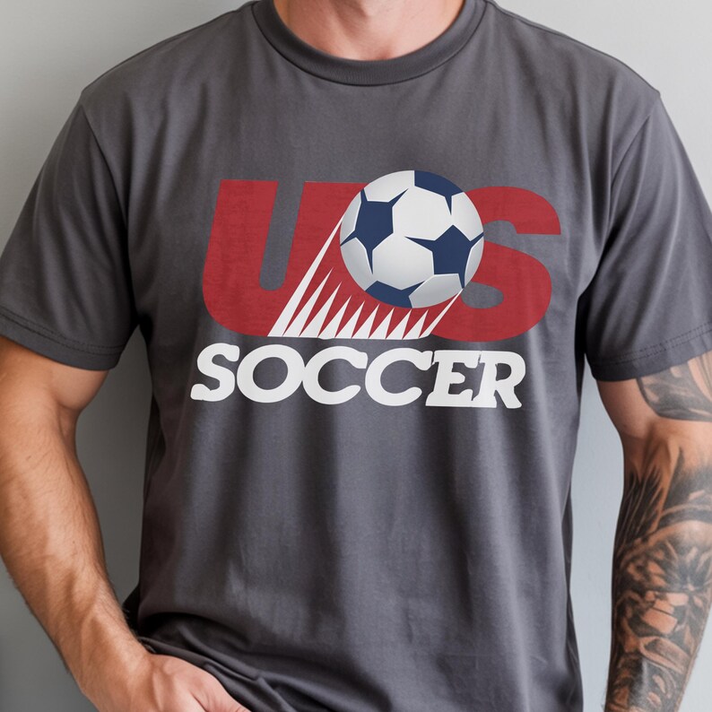 May include: A charcoal grey short-sleeved t-shirt featuring a graphic design with the red letters US, a white and blue soccer ball replacing the letter S, and the word SOCCER printed in large white capital letters underneath the graphic.