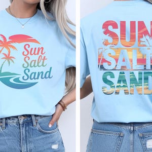 K&ouml;nnte beinhalten: Hellblaues T-Shirt mit tropischem Design. Auf der Vorderseite befinden sich eine Palme und die Worte "Sun Salt Sand" in bunten Farben. Auf der R&uuml;ckseite des Shirts wiederholen sich die Worte "Sun Salt Sand" mit einem Sonnenuntergang und einer Palme.