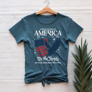 May include: Teal t-shirt with a patriotic design. The graphic includes the text "UNITED STATES OF AMERICA 1776-2026" above a stylized American flag and a soaring eagle. Below, it reads "We the People" and "250-YEAR SEMI-QUINCENTENNIAL."