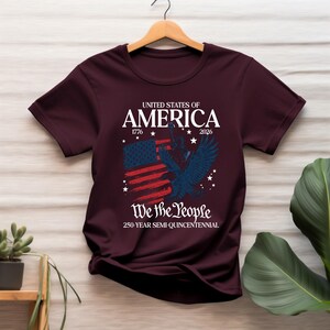 May include: Burgundy t-shirt featuring a patriotic design. The design includes the text "United States of America 1776-2026" above a stylized American flag and a blue eagle. Below the flag is the text "We the People" and "250-Year Semi-Quincentennial."