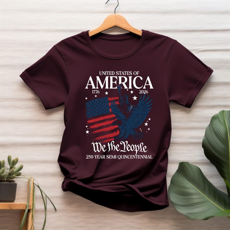 May include: Burgundy t-shirt featuring a patriotic design. The design includes the text "United States of America 1776-2026" above a stylized American flag and a blue eagle. Below the flag is the text "We the People" and "250-Year Semi-Quincentennial."