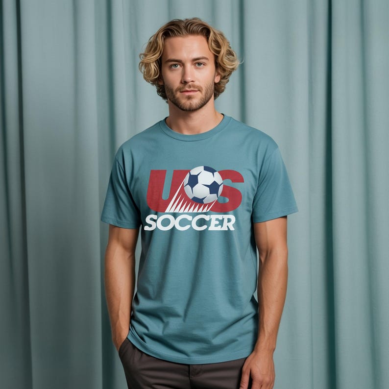 May include: A teal short-sleeved t-shirt featuring a graphic print on the chest. The design includes the red letters US with a white soccer ball positioned in the center, and the word SOCCER printed in white capital letters underneath the ball.