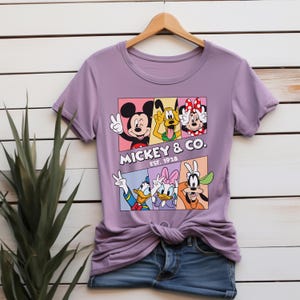 Puede incluir: Una camiseta de color lila con un dise&ntilde;o de Mickey Mouse, el Pato Donald y otros personajes de Disney. El dise&ntilde;o incluye el texto "Mickey & Co. Est. 1928" en blanco. La camiseta est&aacute; colgada en una percha de madera.