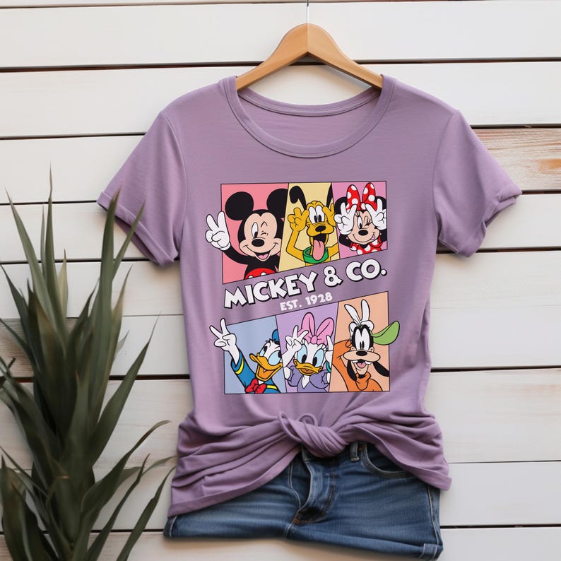 Puede incluir: Una camiseta de color lila con un dise&ntilde;o de Mickey Mouse, el Pato Donald y otros personajes de Disney. El dise&ntilde;o incluye el texto "Mickey & Co. Est. 1928" en blanco. La camiseta est&aacute; colgada en una percha de madera.