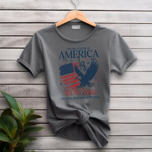 May include: A grey t-shirt with a patriotic design. The shirt features the text "UNITED STATES OF AMERICA" above an illustration of an American flag and an eagle. The text "We the People" is also included, along with the dates 1776 and 2026.